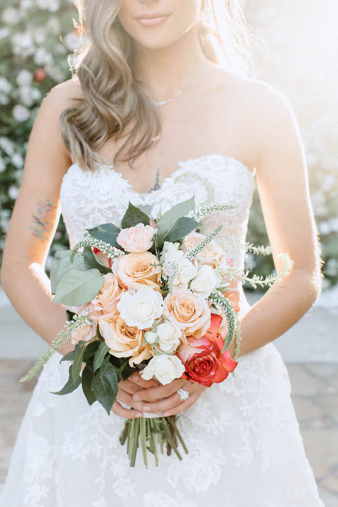 A bride wears a white, lace wedding gown holding her pink, peach, white and red wedding bouquet to display her wedding ring. 