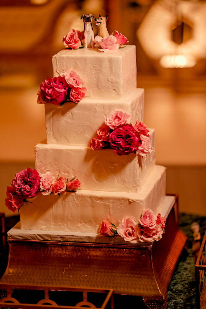 A white wedding cake with pink flowers and clay models of two dogs as cake toppers.