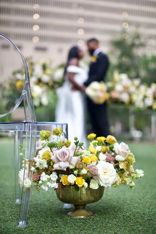 Close-up of yellow, cream and blush florals sitting beside a lucite chair with the couple in the background.