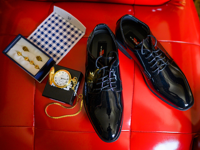 A flat lay of the grooms shoes and accessories on a red leather chair at the wedding venue, Sam Houston Hotel. 