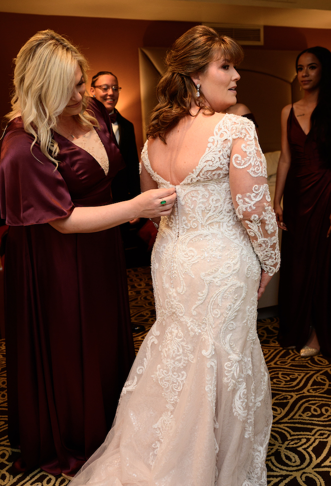 The bride gets buttoned up into her beaded and lace wedding gown for her wedding day by bridesmaid wearing a burgundy gown. 