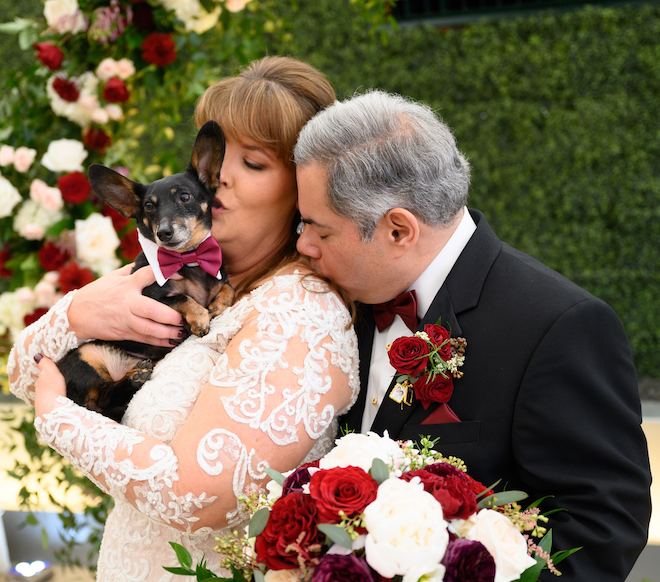 The groom kisses a bride's shoulder while she cuddles a Dachshund wearing a burgundy bow tie. 