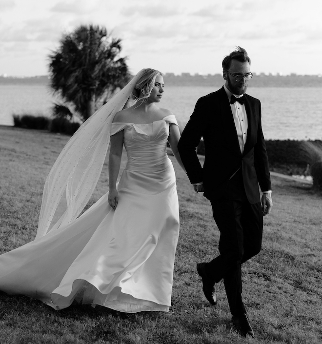 Bride with long veil and off the shoulder wedding gown walks hand in hand with a groom aside a bay at their estate wedding in Sarasota Florida.