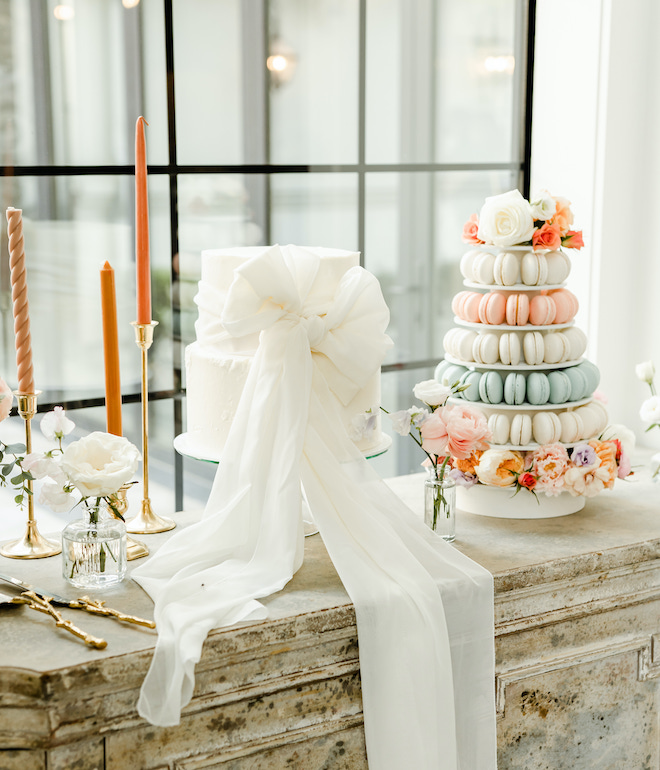 A white wedding cake with a bow and pastel macaroons sit on a table. 