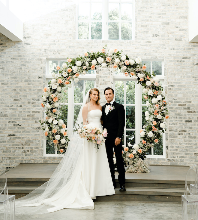 The bride and groom stand in front of a pastel flower arch at the wedding venue, The Peach Orchard in The Woodlands, Texas. 