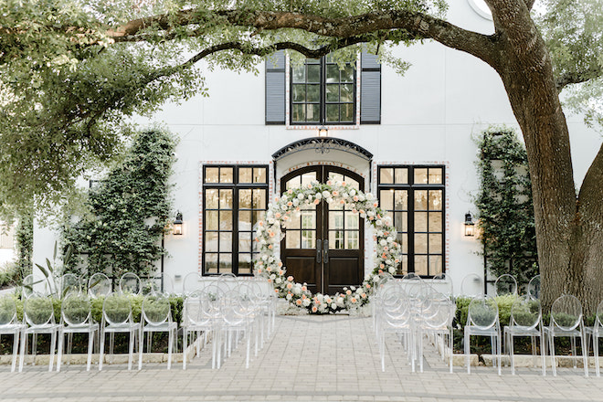A pastel flower arch and acrylic chairs are set up outside the wedding venue, The Peach Orchard. 