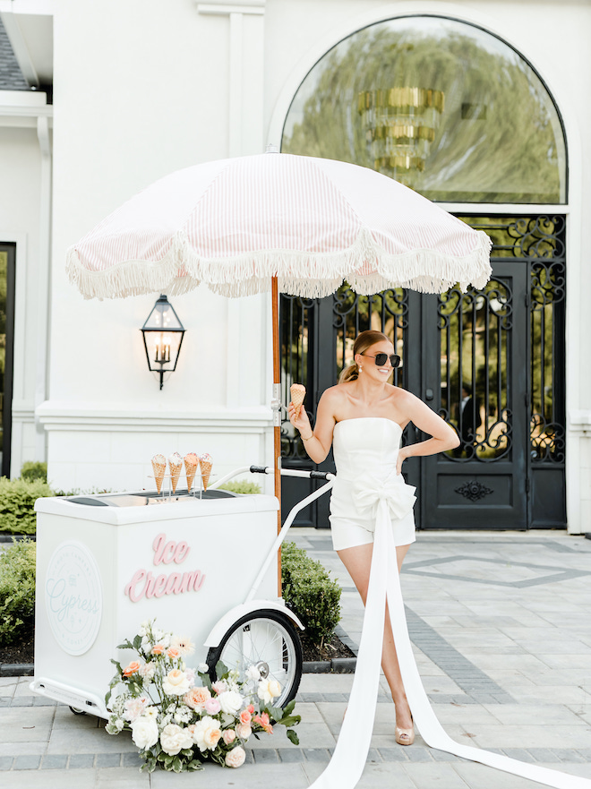 The bride wears sunglasses as she eats an ice cream cone outside her wedding venue. 
