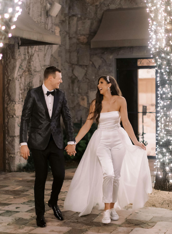 Bride in white jumpsuit holds hands with a smiling groom. 