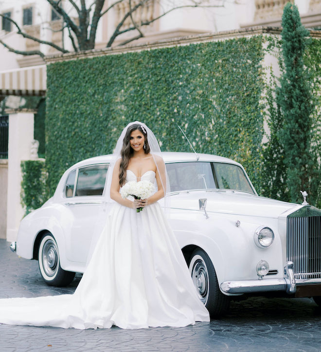 The bride smiles standing beside a white Rolls Royce on her wedding day. 