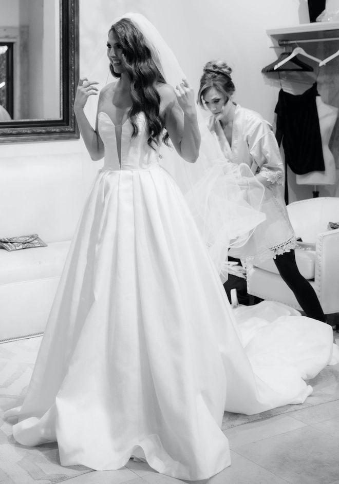 A bride smiles at her reflection in a bridal suite while a women adjusts her long veil at The Bell Tower on 34th in Houston, TX.