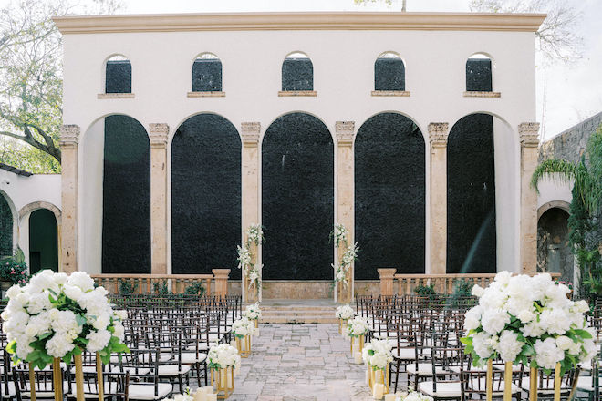 A courtyard with waterwall decorated for a wedding ceremony with white florals and chiavari chairs. 