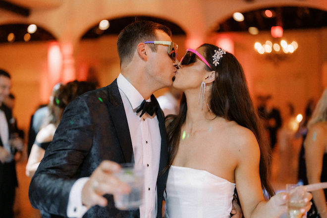 The bride and groom kiss wearing colorful sunglasses on the dance floor at their wedding reception. 