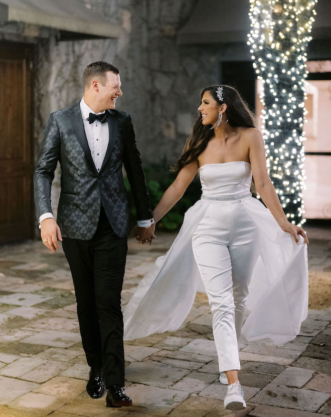 Bride in white jumpsuit and groom hold hands after their New Year's Eve wedding reception.