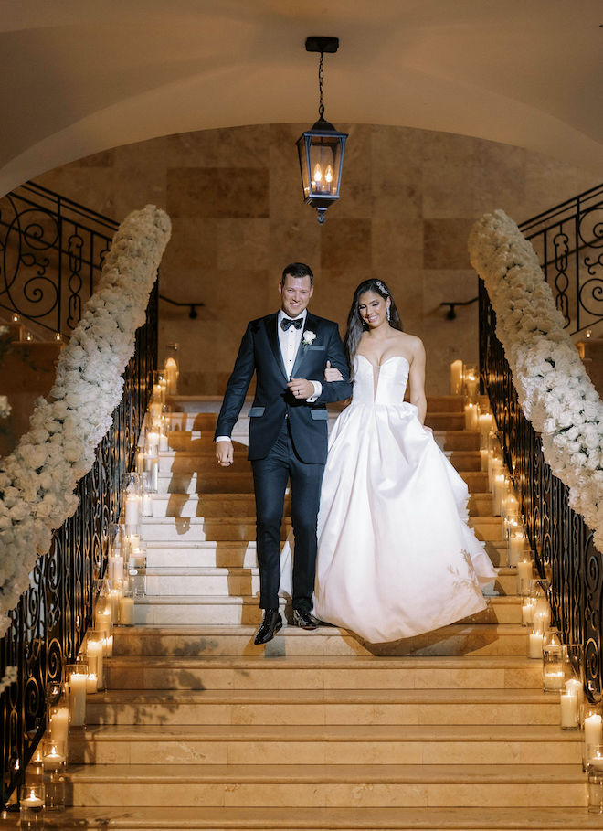 The bride and groom smile as they hold hands while walking down a staircase lined with fresh florals and lit candles. 