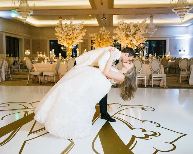 Bride and groom dancing in a ballroom decorated with tall cherry blossom floral arrangements, glowing candles and blush, ivory and gold hues. 