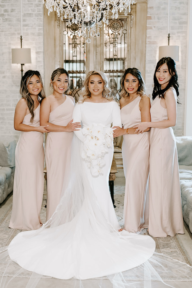 Bride, holding bouquet of lilies, stands in the Soho Suite with bridesmaids wearing blush dresses. 