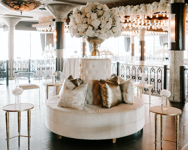 Round lounge furniture decorated with gold and white pillows and a all-white floral centerpiece at a white themed wedding reception at The Astorian.