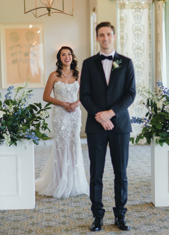 Bride holds her stomach and smiles waiting for her groom to turn around during their first look at their vow renewal at pine forest country club. 