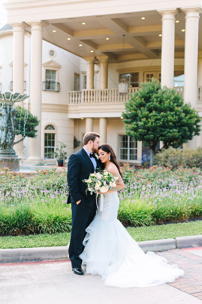 The groom kisses the brides forehead while she is looking down and holding a bouquet in front of the venue.