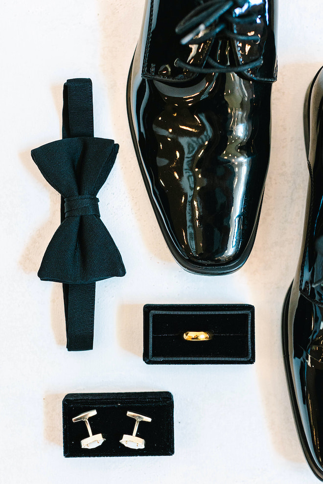 The groom's black bowtie, black shiny shoes, gold ring and cufflinks arranged for a photo.