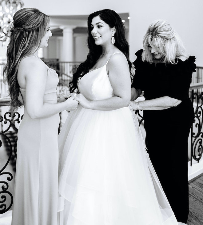The bride holding a bridesmaids hand while her mother helps her with her dress. 