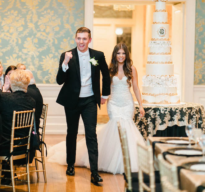Groom raises his fist and smiles while holding hands with a bride in a fit and flare wedding gown as they walk into their black tie wedding reception.