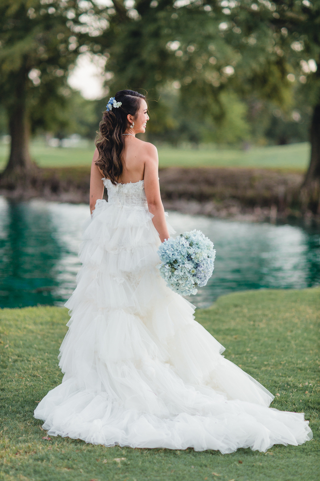 The back of a bride wearing a ruffled corset style wedding gown from Belle Ame Bridal holding a blue and purple bouquet of hydrangeas on the golf green of pine forest country club.