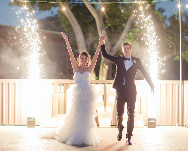 Bride and groom hold hands and walk forward while two sparklers go off on either side of them with a lit sign behind them that reads, "Love" after their vow renewal at Pine Forest Country Club. 