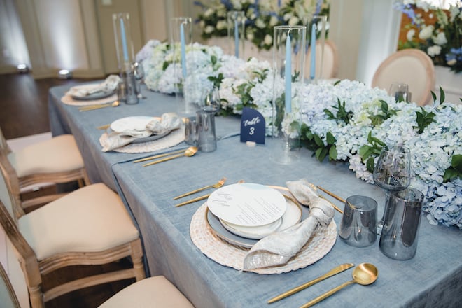 A family style tablesetting with rattan chargers, gold napkins, navy placecards and blue florals in a ballroom at Pine Forest Country Club. 