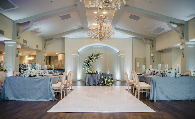 The Legacy Ballroom at Pine Forest Country Club decorated with two family style tables, blue tableclothes, blue hydrangea, a white dance floor and lilac tapered candles. 