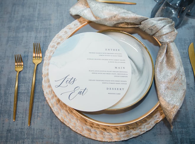 A placesetting that reads, "Let's Eat" and "Entree" "Main" and "Dessert" in cursive font a top a blue dinner plate, gold napkins and gold utensils at a vow renewal in the Legacy Ballroom at Pine Forest Country Club.