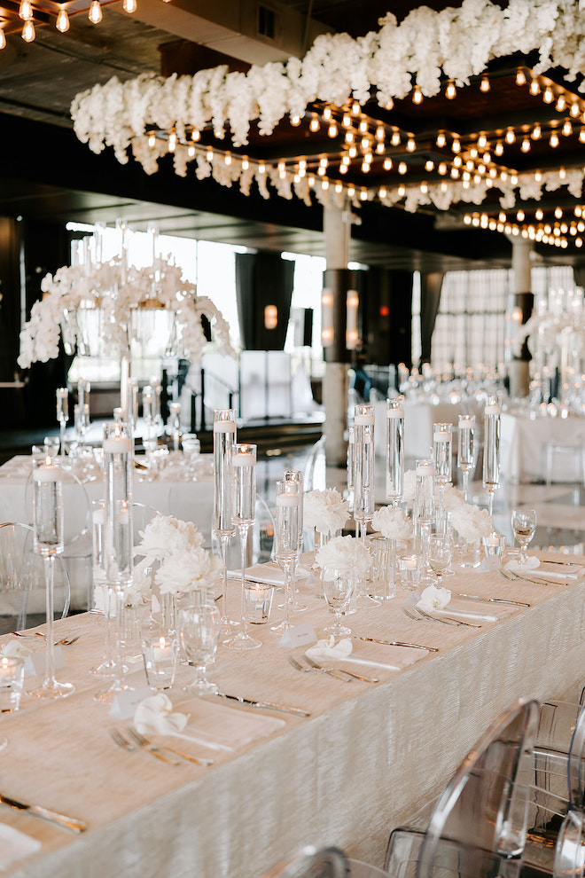 A family style reception table set with blooming white florals, lit votive candles and ivory tablecloth and ghost chairs in the ballroom of The Astorian. 