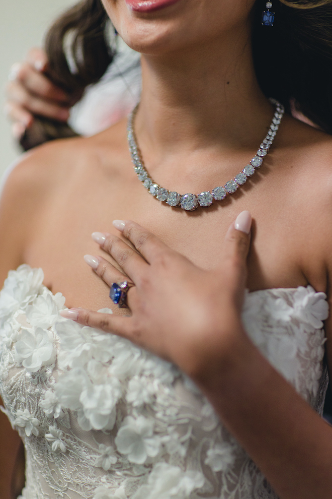 A bride wearing a diamond necklace from Privosa, a floral applique wedding dress from Belle Ame Bridal Houston and an Blue Sapphire Wedding Ring while her hair is being curled by a makeup artist.