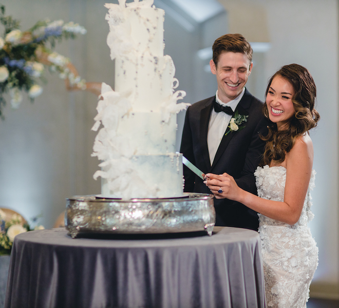 bride and groom holding a cake knife next to a four tier white wedding cake with light blue and silver accents at their reception at pine forest country club in houston, texas. 