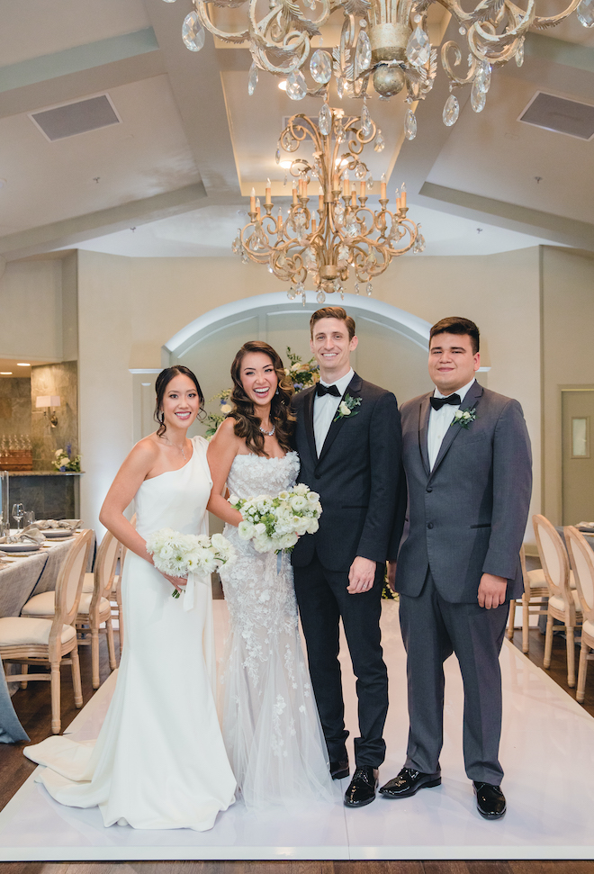 Bride and groom smile with a bridesmaid and groomsmen at their vow renewal and reception in a ballroom with gold chandeliers.