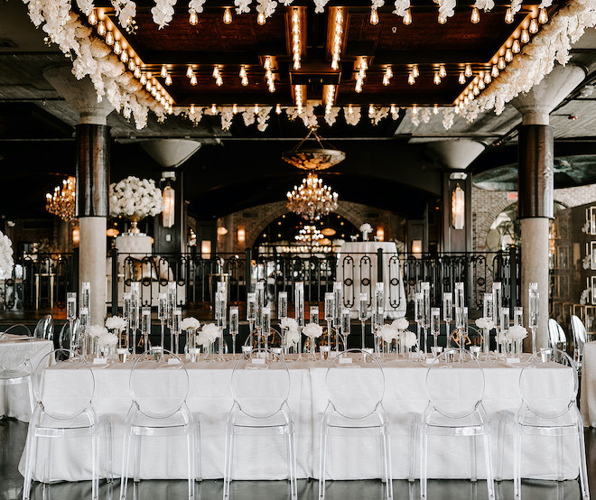 Family style table decorated with candles, white floral centerpieces and ghost chairs at a wedding reception in the Astorian's ballroom. 