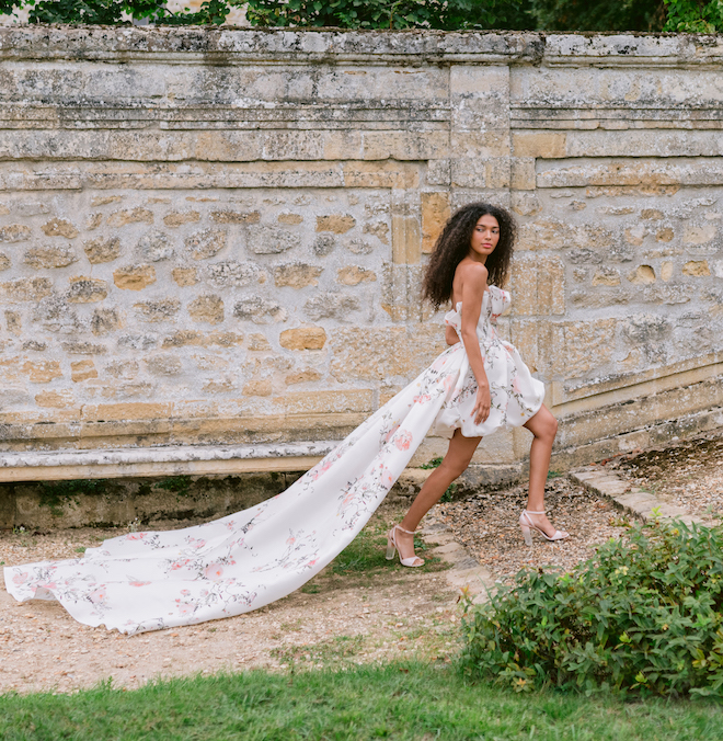 Model wearing a short floral dress with a long cape trailing behind her. 