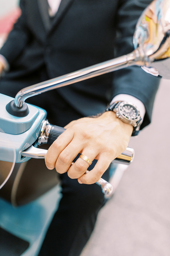 Man wearing a gold wedding band and luxury watch while riding a blue vespa motorcycle at a wedding editorial in Texas. 