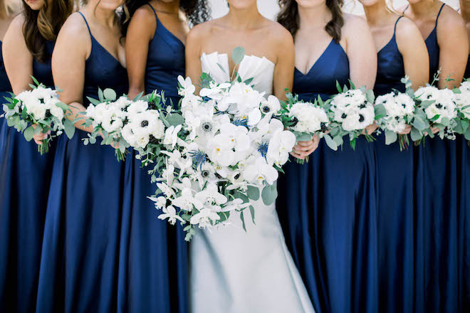 The bride standing in the middle of her bridesmaids dressed in blue gowns, all of them holding their bouquets of white and navy hues.