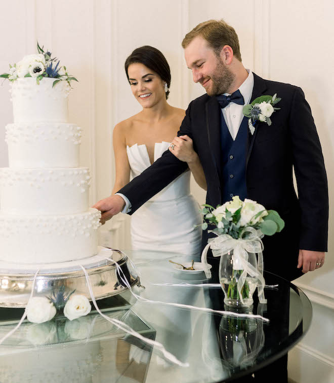 The bride holds the grooms arm while he cuts the four-tiered white wedding cake.