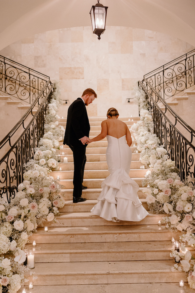 Newly married bride and groom walk hand in hand on a staircase lined with lit candles and blush and white florals at their wedding reception at the Bell Tower on 34th. 