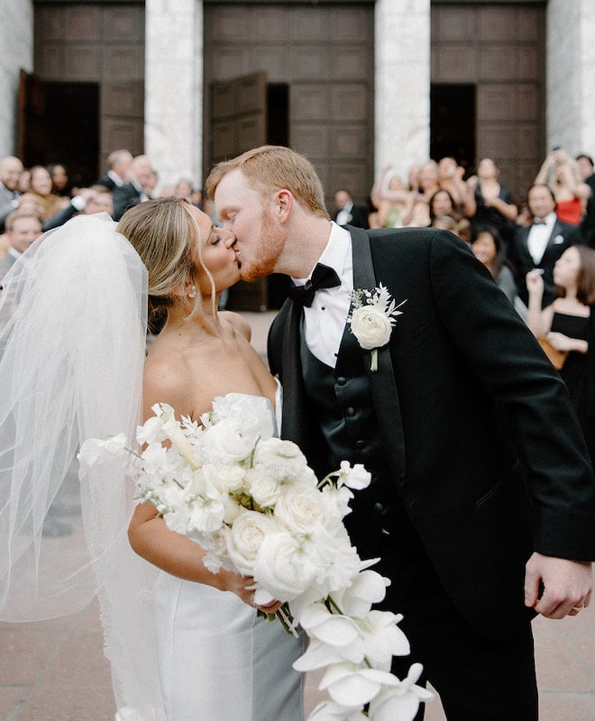 Bride holding white orchid and rose bouquet kisses a groom in a black suit after a church ceremony in Houston, Texas. 