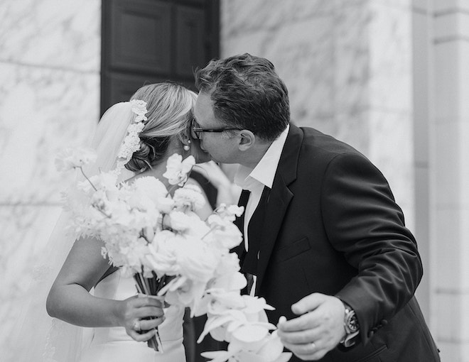 A bride holds a white bouquet of orchids while her father kisses her cheek before her church ceremony. 