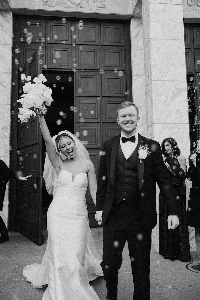 Bride raises her arm while holding hands with a groom while guests blow bubbles after their church ceremony in Houston. 