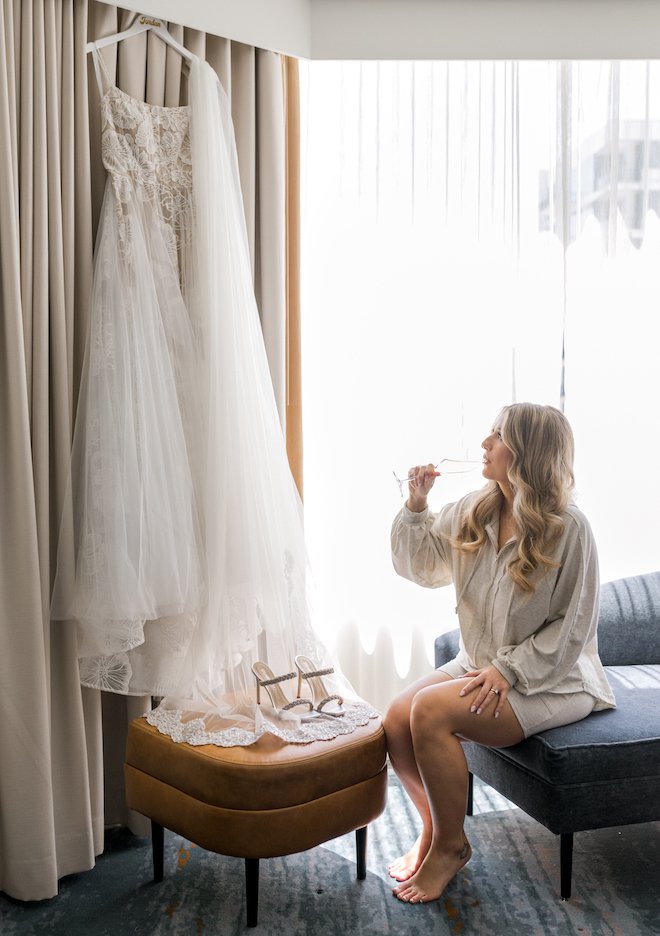 The bride sipping champagne as she looks up at her hanging wedding gown. 