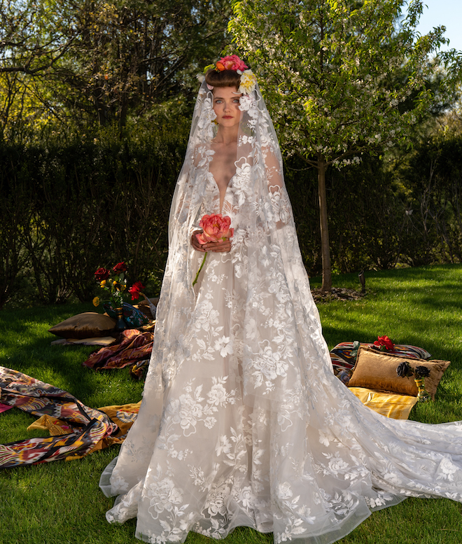 Model wearing a lace bridal gown with a matching veil and holding an orange flower.