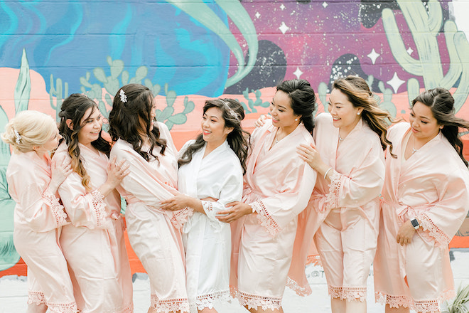 The bridesmaids hugging the bride in their blush pink robes with a painted mural on the wall building behind them.