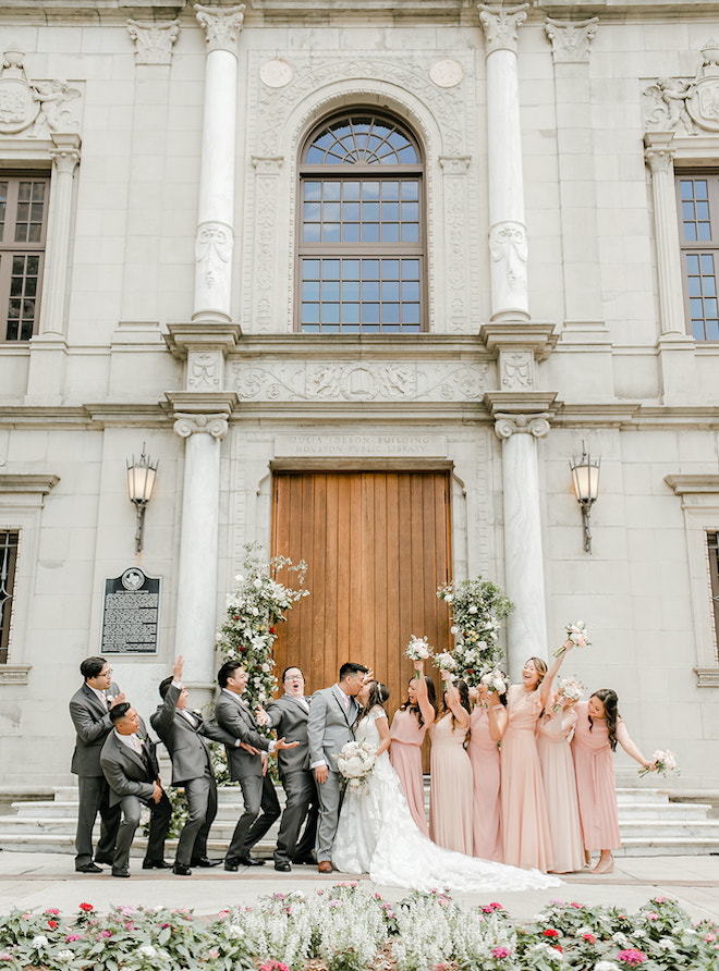 The bride and groom kissing in front of the library as the wedding party cheers on either side of them.