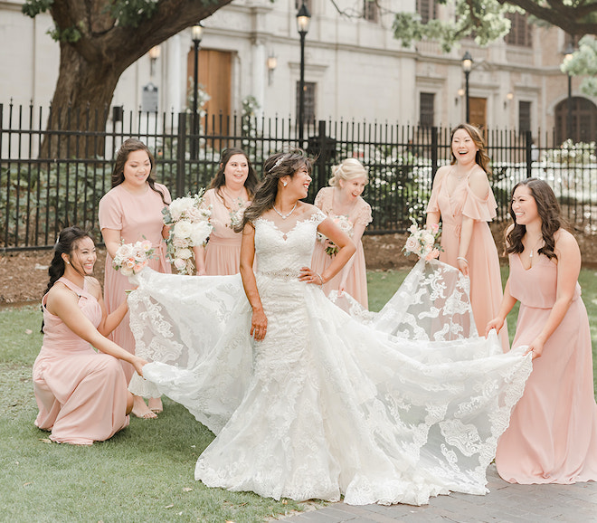 The bridesmaids holding the bottom of the brides gown as they all smile at each other. 