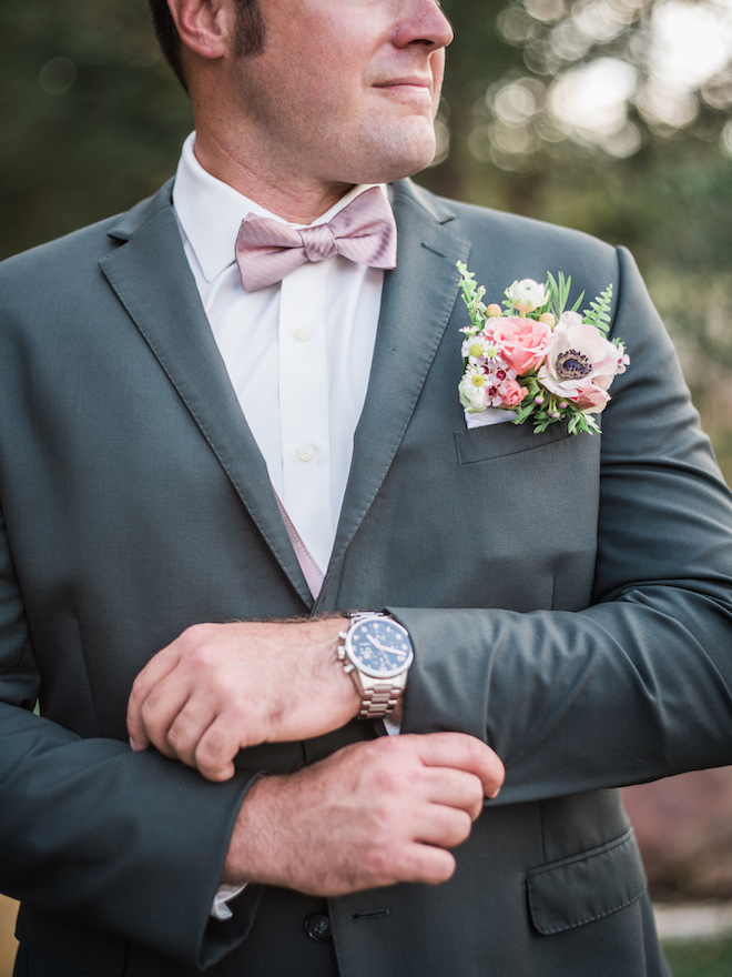 The groom in a gray suit with a pink bow tie with florals coming out of his pocket square.